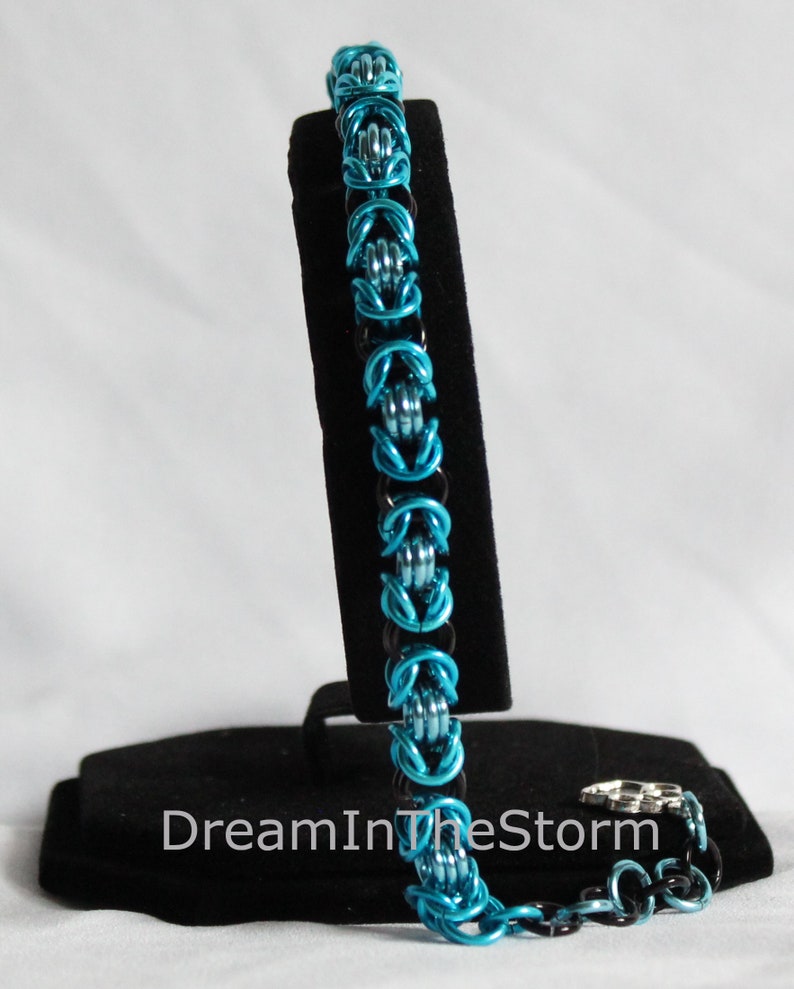 Puede incluir: Un brazalete de malla de cadena azul y negro con acabado plateado. El brazalete se muestra en un soporte negro con el texto "DreamInTheStorm".