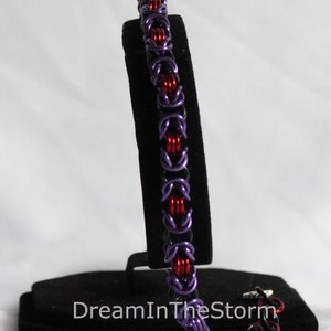 Puede incluir: Un brazalete de malla de color morado y rojo sobre un soporte negro. El brazalete est&aacute; hecho de peque&ntilde;os anillos de metal. El texto "DreamInTheStorm" es visible en el soporte.