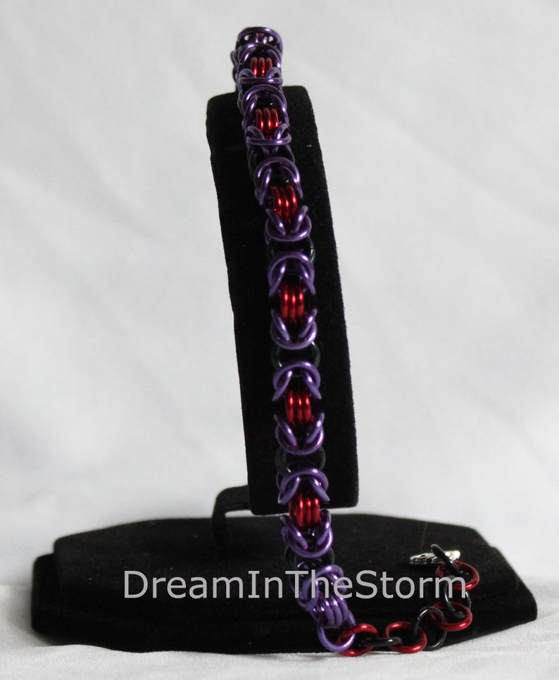 Puede incluir: Un brazalete de malla de color morado y rojo sobre un soporte negro. El brazalete est&aacute; hecho de peque&ntilde;os anillos de metal. El texto "DreamInTheStorm" es visible en el soporte.