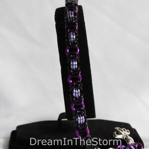 Puede incluir: Un brazalete de malla de cadena negro y morado con un cierre plateado. El brazalete se muestra en un soporte de terciopelo negro.