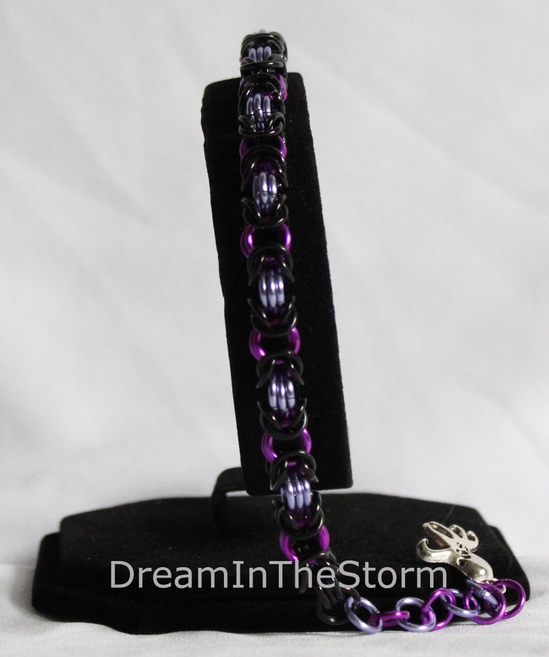 Puede incluir: Un brazalete de malla de cadena negro y morado con un cierre plateado. El brazalete se muestra en un soporte de terciopelo negro.