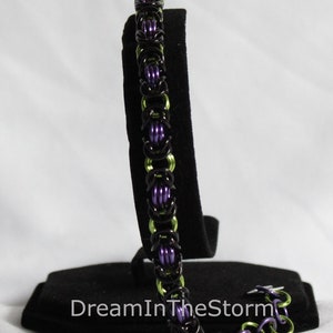 Puede incluir: Un brazalete de malla de cadena negro, verde y morado con un cierre. El brazalete se muestra en un soporte de terciopelo negro.