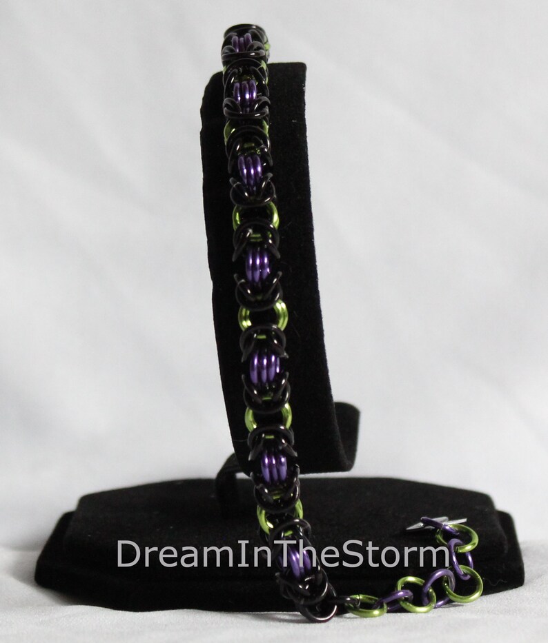 Puede incluir: Un brazalete de malla de cadena negro, verde y morado con un cierre. El brazalete se muestra en un soporte de terciopelo negro.