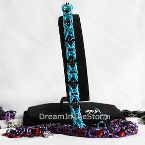Puede incluir: Tres pulseras de malla de cadena en negro, azul verdoso y morado con detalles plateados. Las pulseras se muestran en un soporte negro con el texto "DreamInTheStorm".
