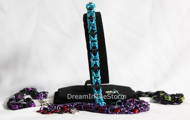 Puede incluir: Tres pulseras de malla de cadena en negro, azul verdoso y morado con detalles plateados. Las pulseras se muestran en un soporte negro con el texto "DreamInTheStorm".
