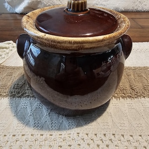 May include: A round, dark brown ceramic bean pot with a matching lid. The pot features a glossy finish, two side handles, and a textured, lighter tan rim around the top opening. The lid has a small, ridged knob handle at the center.
