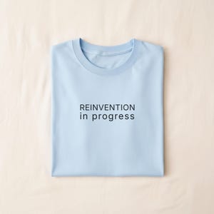 May include: A light blue t-shirt with the text "REINVENTION in progress" in black. The shirt is folded neatly on a beige surface.
