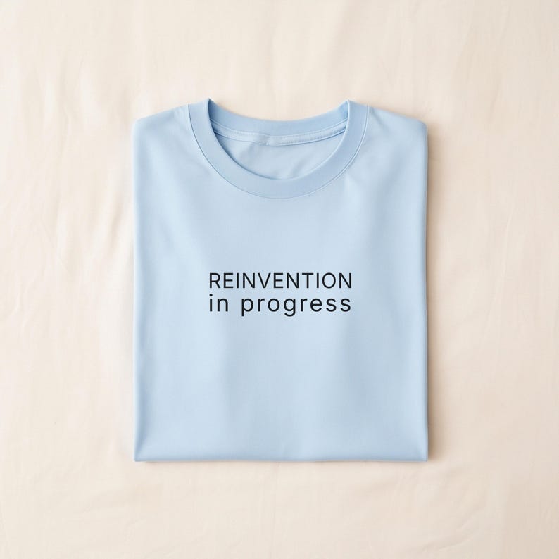 May include: A light blue t-shirt with the text "REINVENTION in progress" in black. The shirt is folded neatly on a beige surface.