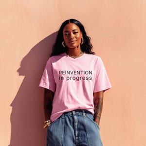 May include: A pink t-shirt with the text "REINVENTION in progress" in black. The shirt is paired with blue denim jeans. The person is wearing gold jewelry, including earrings and bracelets. The background is a solid pink wall.
