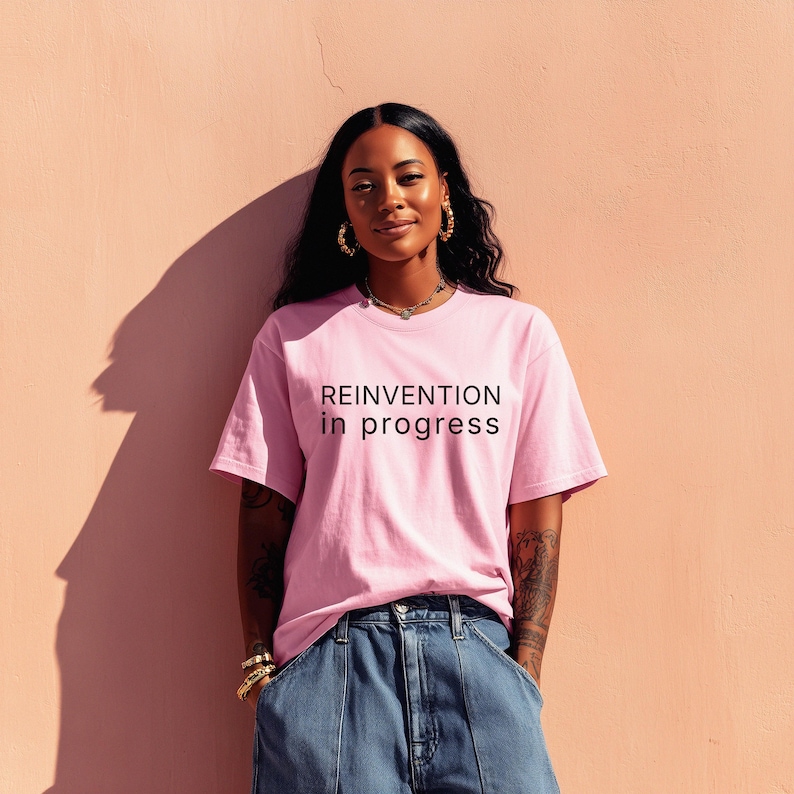 May include: A pink t-shirt with the text "REINVENTION in progress" in black. The shirt is paired with blue denim jeans. The person is wearing gold jewelry, including earrings and bracelets. The background is a solid pink wall.
