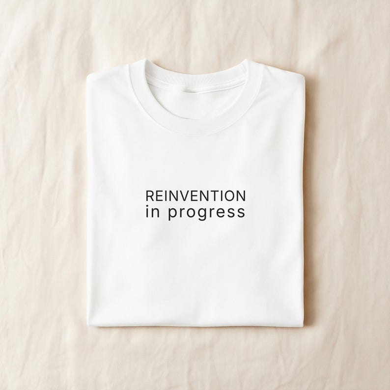May include: A folded white t-shirt with the text "REINVENTION in progress" printed in black. The shirt is made of a soft material and is neatly folded, suggesting it is new or freshly laundered.