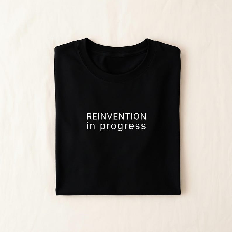 May include: Black t-shirt with the white text "REINVENTION in progress". The shirt is folded neatly, showcasing the design. The fabric appears to be a soft cotton material, suitable for casual wear.