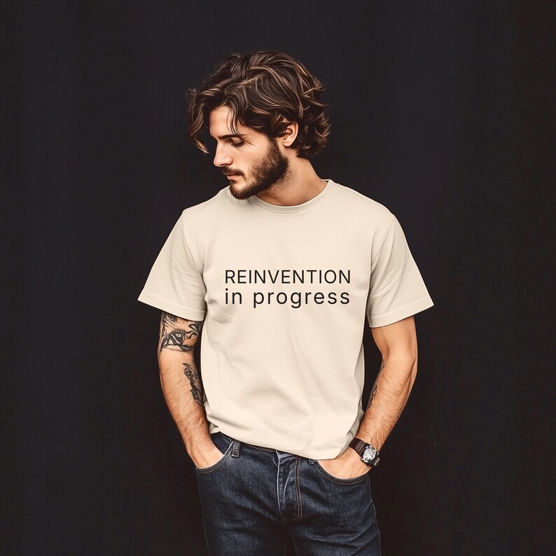 May include: Cream-colored t-shirt with the text "REINVENTION in progress" in black. The shirt is paired with blue jeans. The person is wearing a watch on their left wrist.