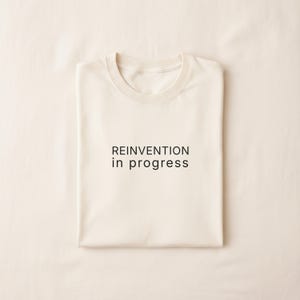 May include: Cream-colored t-shirt with the phrase "REINVENTION in progress" printed in black. The shirt is neatly folded, showcasing the round neckline and overall design. The fabric appears soft and comfortable.