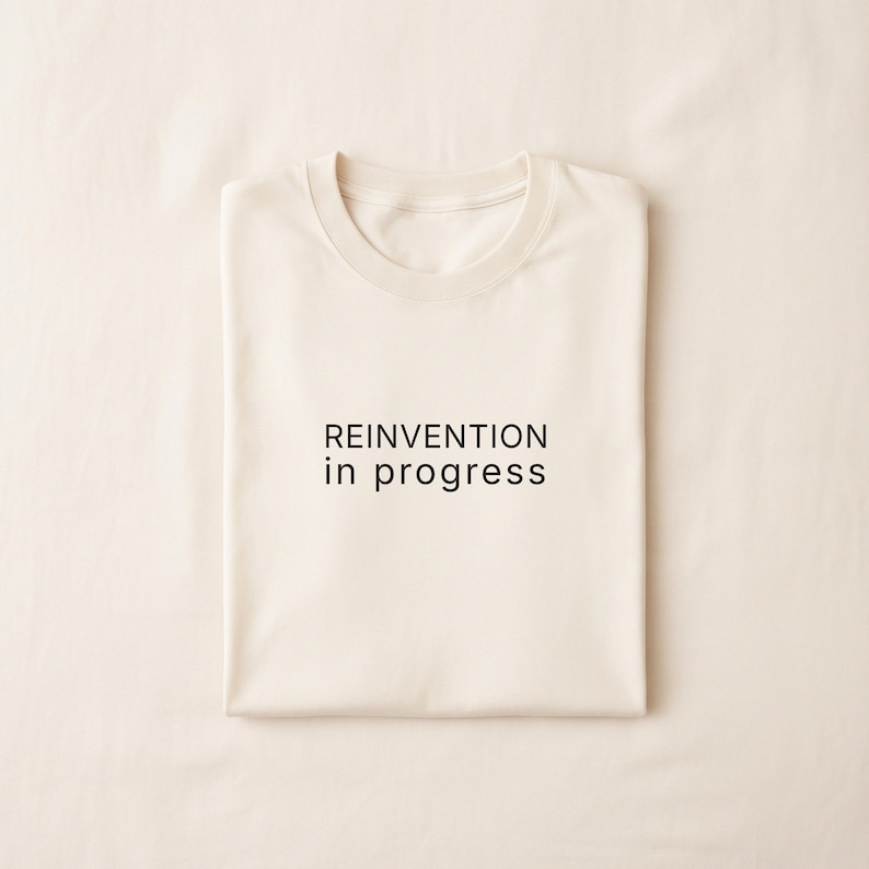 May include: Cream-colored t-shirt with the phrase "REINVENTION in progress" printed in black. The shirt is neatly folded, showcasing the round neckline and overall design. The fabric appears soft and comfortable.