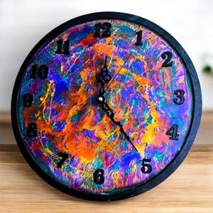 Pode incluir: Relógio de parede redondo com moldura preta e design abstrato colorido. O mostrador do relógio apresenta uma vibrante mistura de tons laranja, azul e roxo, com algarismos e ponteiros pretos. O relógio é um item decorativo.
