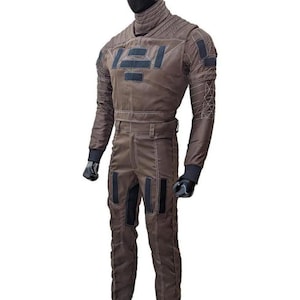 Op de afbeelding: Een bruin jumpsuit met zwarte accenten, getoond op een mannequin. Het pak heeft een hoge kraag, meerdere zakken en een riem. De mouwen hebben een geometrisch patroon op de bovenarmen. De mannequin draagt zwarte handschoenen en laarzen.