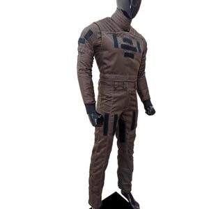 Op de afbeelding: Een bruin jumpsuit met zwarte accenten, gepresenteerd op een mannequin. Het pak heeft een hoge kraag, lange mouwen en meerdere rechthoekige patches. Zwarte handschoenen en laarzen maken de outfit compleet. De jumpsuit lijkt gemaakt van duurzaam materiaal.