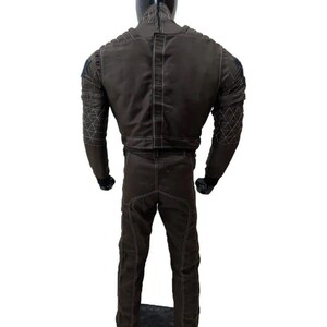 Op de afbeelding: Bruin jumpsuit met witte stiksels en gewatteerde details op de schouders en mouwen. Het pak heeft een hoge kraag en een rits op de rug. De mannequin draagt zwarte handschoenen.