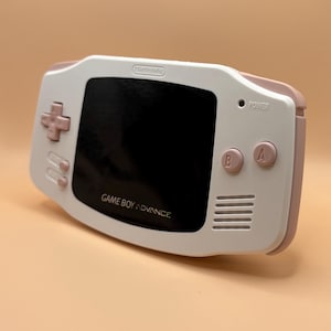 Könnte beinhalten: Ein weiß-rosa Game Boy Advance Handheld-Konsolen. Die Konsole hat einen schwarzen Bildschirm, ein rosa Steuerkreuz und rosa A- und B-Tasten. Der Text "GAME BOY ADVANCE" ist unter dem Bildschirm aufgedruckt.