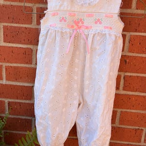 May include: White eyelet baby romper with a lace collar and pink ribbon accents. The romper features a smocked bodice with pink floral embroidery and a delicate bow. The garment is hanging on a metal hanger against a brick wall.