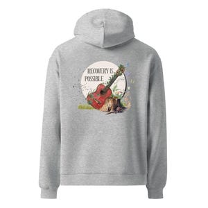 Puede incluir: Una sudadera con capucha gris claro con un gráfico en la espalda. El gráfico presenta una guitarra roja apoyada en un tocón de árbol, con las palabras "RECOVERY IS POSSIBLE" en un círculo blanco. Mariposas y follaje rodean la guitarra.