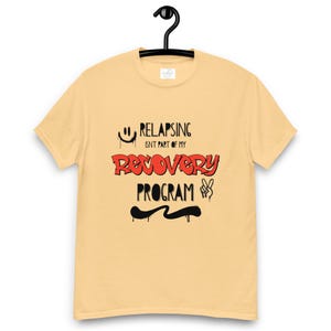 May include: A light yellow t-shirt with the text "Relapsing isn't part of my Recovery Program" in black and red. The shirt is hanging on a black hanger.
