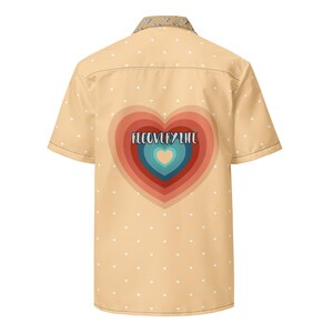 May include: Short-sleeved button-up shirt in a light tan color, adorned with small white hearts. The back features a large, layered heart design in shades of red, orange, and blue, with the text "RECOVERY LIFE" in the center. The collar has a patterned design.