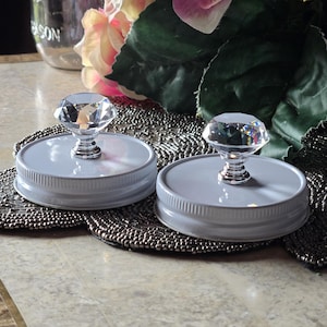 Peut inclure: Deux couvercles de bocaux gris ornés de boutons en cristal taillé. Les boutons réfléchissent la lumière. Les couvercles reposent sur un dessous de plat perlé, avec des fleurs artificielles roses et vertes en arrière-plan. Un bocal Mason est également visible.