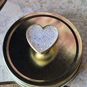 Könnte beinhalten: Goldfarbener Deckel für ein Einmachglas mit herzförmigem Griff. Das Herz ist mit schillernden, kristallartigen Elementen gefüllt. Der runde Deckel hat eine metallische Oberfläche, ideal für Heimdekoration oder Bastelprojekte.