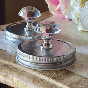 Puede incluir: Dos tapas decorativas para tarros con pomos de cristal facetado. Las tapas son de metal plateado con un borde acanalado. Los pomos de cristal son transparentes y reflejan la luz. El fondo incluye flores rosas y blancas.