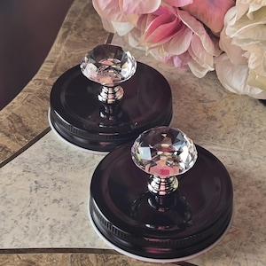 May include: Two black jar lids with clear crystal-like knobs. The knobs have a silver base. The lids are on a patterned surface, with pink and white flowers in the background. The jar lids are likely for decorative purposes.