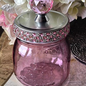 May include: A pink glass jar with a decorative lid. The lid features a silver rim adorned with pink rhinestones and a faceted pink crystal knob. The jar is filled with a pink liquid and has embossed detailing.