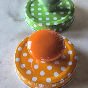 May include: Two circular metal jar lids with white polka dots. One lid is bright green with a matching green knob handle, and the other is vibrant orange with a matching orange knob handle. Both lids feature a ridged edge and a smooth finish.