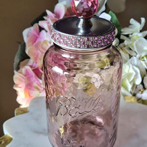 Peut inclure: Un bocal en verre rose Ball Ideal avec un couvercle argenté. Le couvercle présente une bande de petits strass roses et un bouton en cristal rose à facettes sur le dessus. Le verre a une surface texturée et le texte Ball et IDEAL en relief.