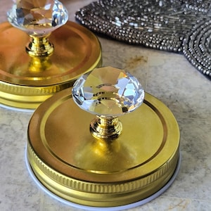 May include: Two gold-coloured jar lids with clear, faceted crystal knobs. The lids have a white rubber seal. The knobs are attached to the lids with a gold-coloured base. The background includes a beaded item and pink and white flowers.