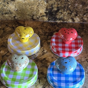 Puede incluir: Cuatro tapas de tarro con pomos en forma de corazón. Cada tapa tiene un color y un patrón diferente: amarillo con cuadros amarillos y blancos, rojo con cuadros rojos y blancos, verde con cuadros verdes y blancos y azul con cuadros azules y blancos.