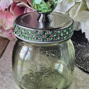 May include: A decorative glass jar with a silver lid and a faceted green crystal knob. The lid is adorned with a band of green and clear rhinestones. The jar is clear with a subtle green tint.