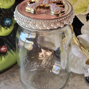 Op de afbeelding: Een heldere glazen Mason jar met een decoratief deksel. Het deksel is versierd met een gouden strik versierd met imitatieparels en strass-steentjes. De pot is leeg en klaar om gevuld te worden.