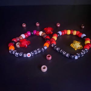May include: Two circular beaded bracelets featuring black square letter beads spelling REMUS LUPIN and SIRIUS BLACK. The bracelets include colorful round plastic beads in red, orange, yellow, and brown, along with red and yellow star-shaped charms.