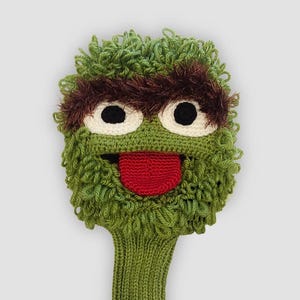 May include: A green and brown crocheted golf club head cover shaped like a cartoon character with large white eyes, a red tongue, and shaggy green fur. The cover is designed to protect golf clubs.
