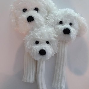 May include: Three white, fluffy dog headcovers for golf clubs. Each cover features a plush, white exterior with black eyes and a black nose. The covers have long, ribbed, white knit sleeves.