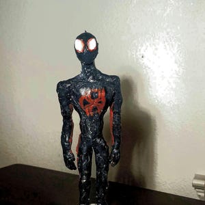May include: A handmade figurine of a superhero in a black suit with red accents. The figure has glowing white eyes and a red spider emblem on the chest. It stands on a white base.