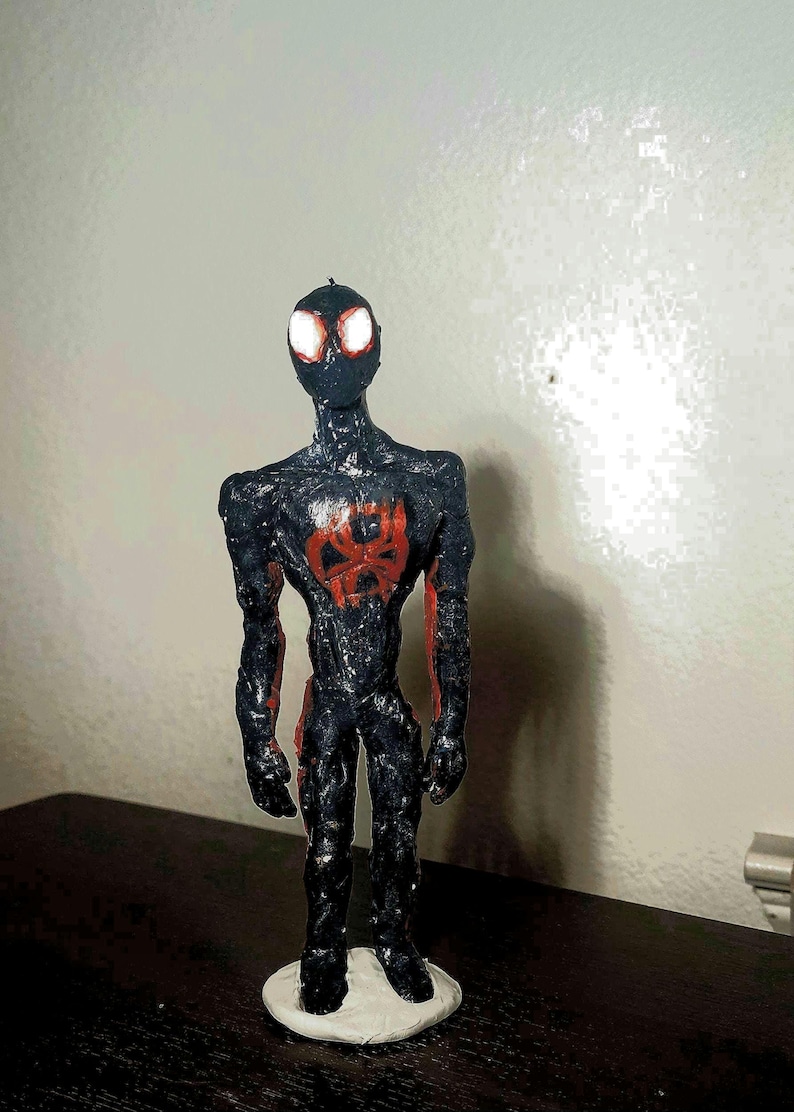 May include: A handmade figurine of a superhero in a black suit with red accents. The figure has glowing white eyes and a red spider emblem on the chest. It stands on a white base.