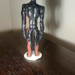 May include: A handmade sculpture of a humanoid figure. The figure is painted black with red accents on the legs and torso. It stands on a white circular base. The sculpture is on a dark brown surface.