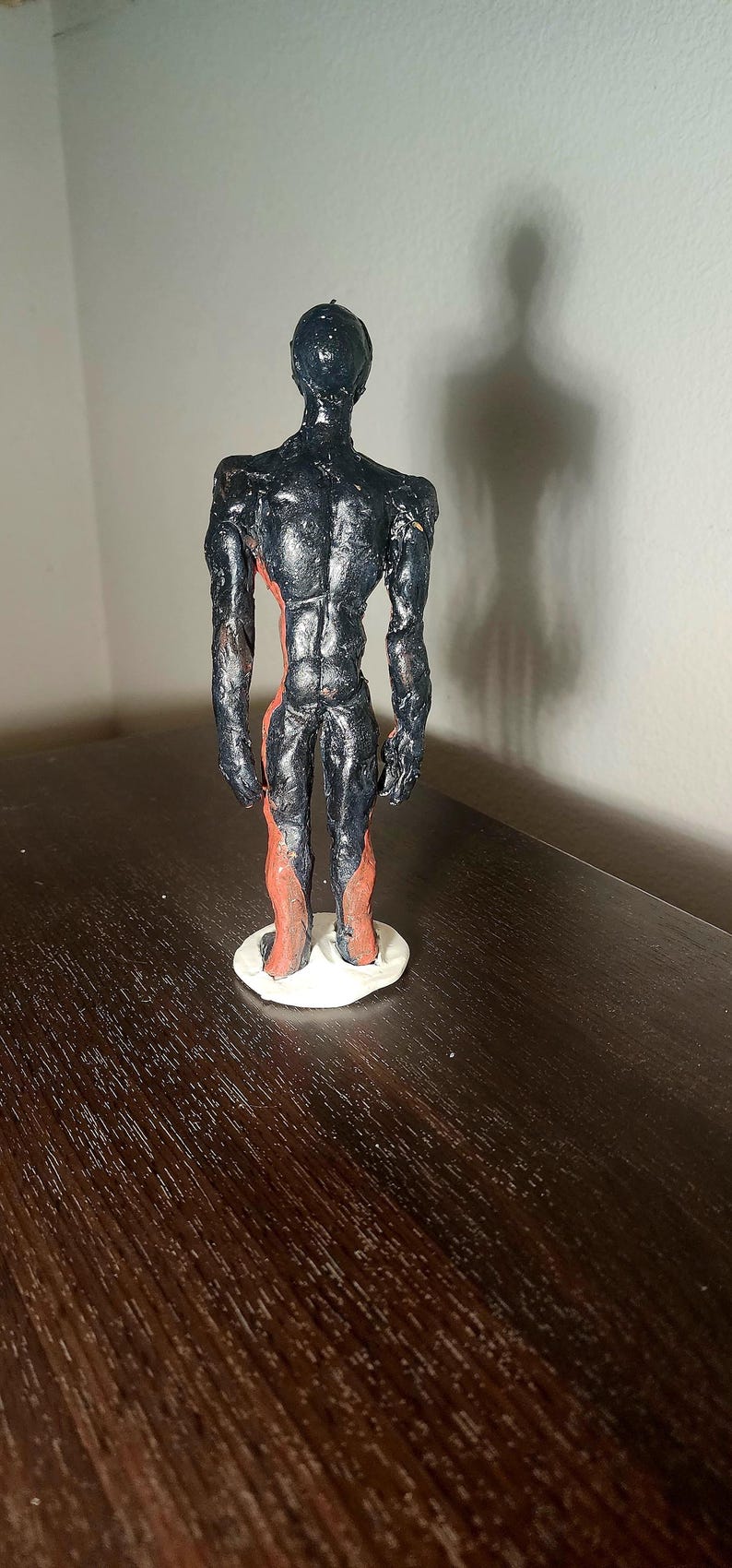 May include: A handmade sculpture of a humanoid figure. The figure is painted black with red accents on the legs and torso. It stands on a white circular base. The sculpture is on a dark brown surface.