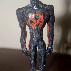 May include: A handmade figurine of a superhero, featuring a black and red color scheme. The figure has a textured appearance, with a red spider emblem on the chest and white eyes. It stands on a white base.