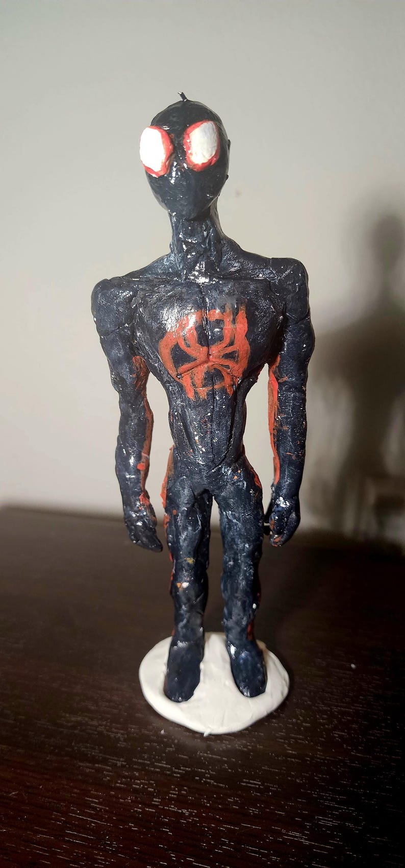 May include: A handmade figurine of a superhero, featuring a black and red color scheme. The figure has a textured appearance, with a red spider emblem on the chest and white eyes. It stands on a white base.