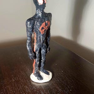 May include: A handmade figurine of a superhero, featuring a black and red color scheme. The figure stands on a white base, with a detailed design of the character's suit. The figure is posed in a standing position.