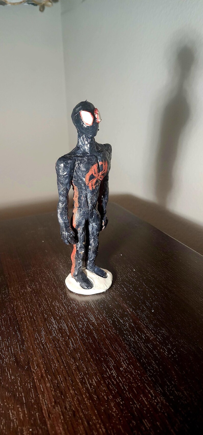 May include: A handmade figurine of a superhero, featuring a black and red color scheme. The figure stands on a white base, with a detailed design of the character's suit. The figure is posed in a standing position.
